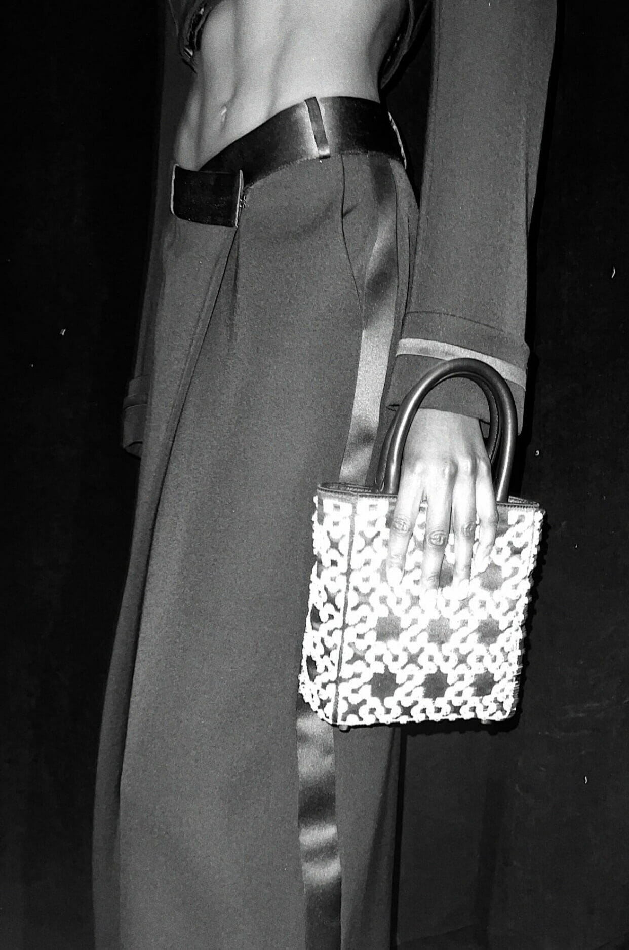 Black-and-white image of a model holding a structured SIMKHAI handbag styled with tailored trousers and a cropped top.