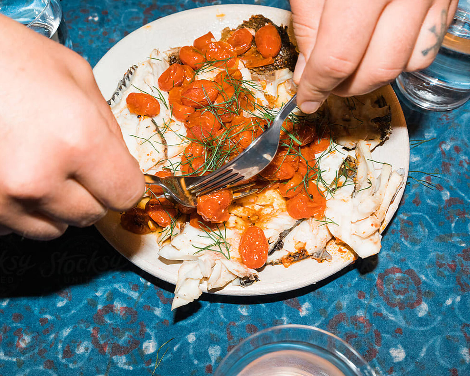 shot of hands cutting into a plate of roasted white fish topped with blistered cherry tomatoes and fresh dill on a blue patterned tablecloth